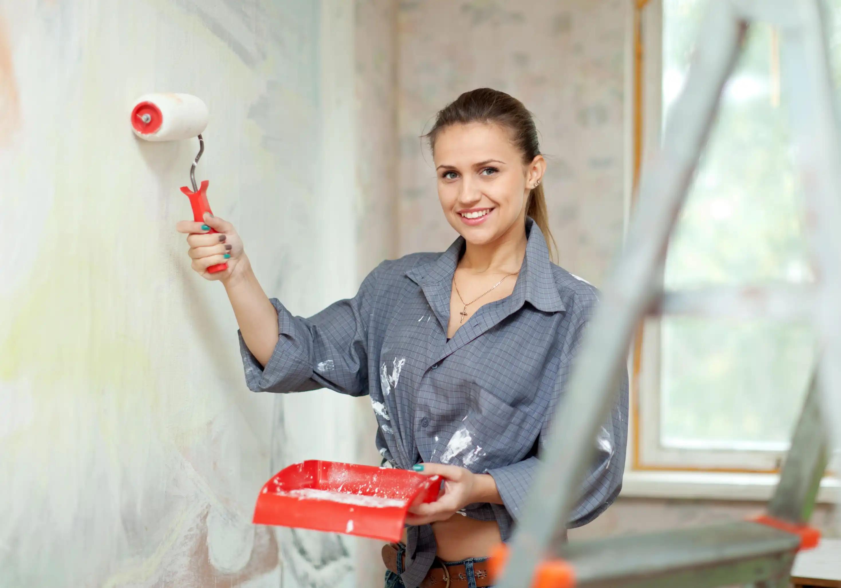Woman painting a wall with a roller during home renovation, holding a paint tray and smiling indoors